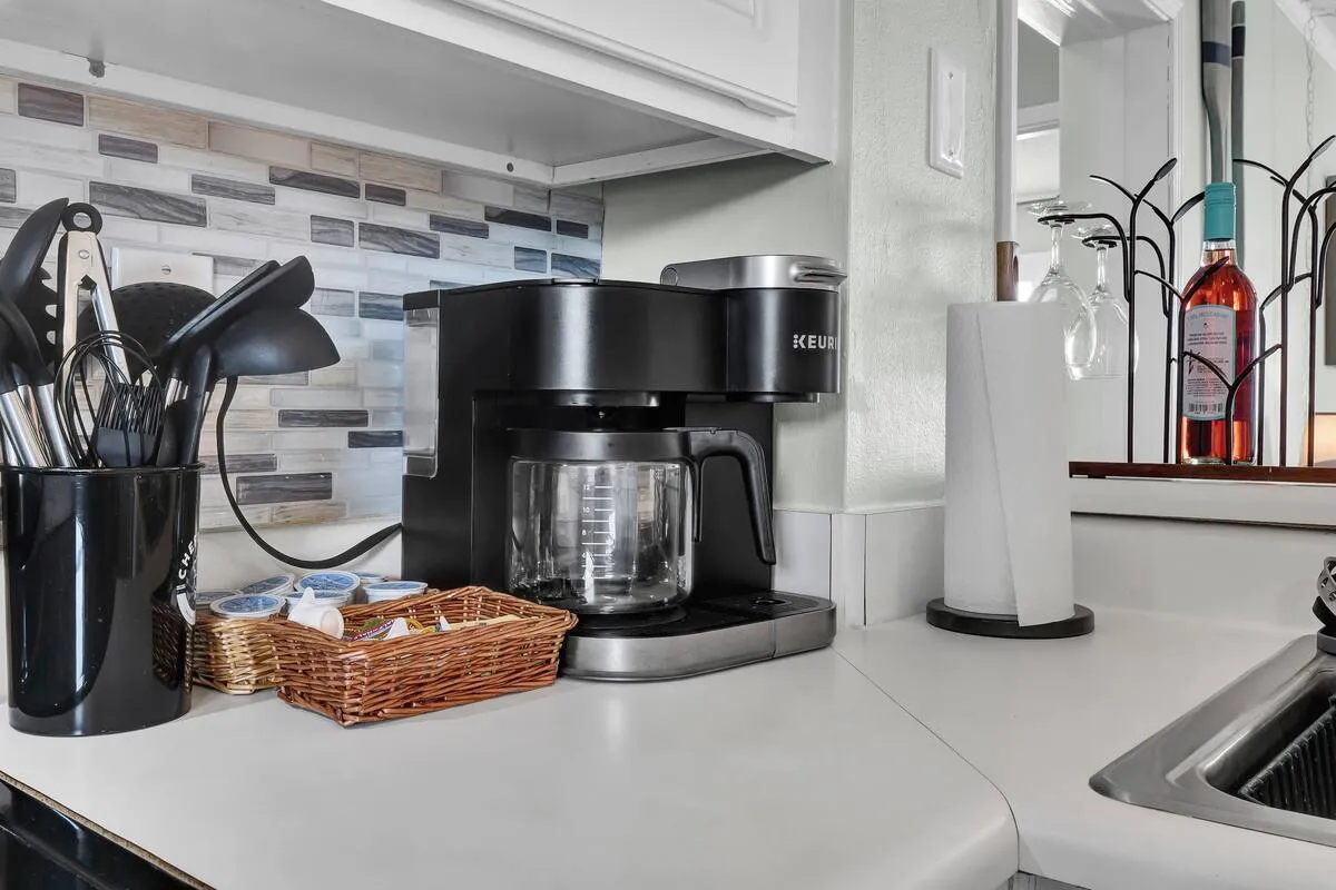 A close-up of a coffee maker, utensil holder, basket of coffee pods, and a wine bottle on the kitchen counter. The scene evokes cozy morning routines and intimate evening gatherings.