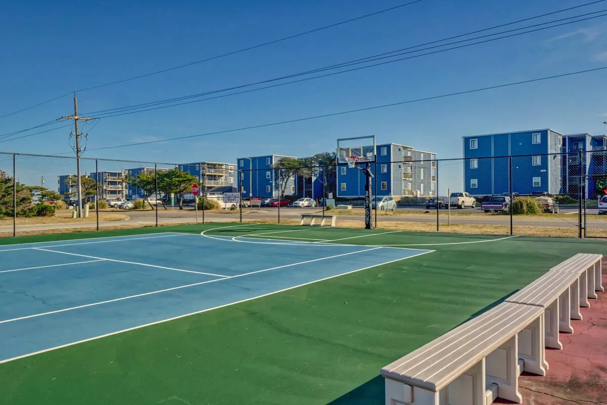 A bright basketball court surrounded by benches and nearby buildings, inviting residents and visitors to engage in friendly games, exercise, or casual gatherings. The space clearly connects to the wider community and encourages outdoor activity.