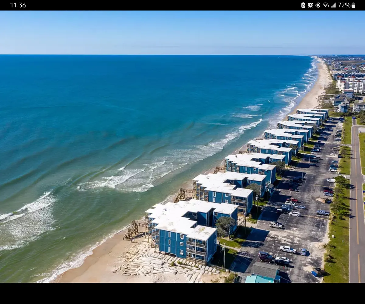 A scenic exterior view of beachfront properties with the ocean, clear sky, parked cars, and a coastal path. The image conveys immediate proximity to the beach and the inviting environment just outside the condo.