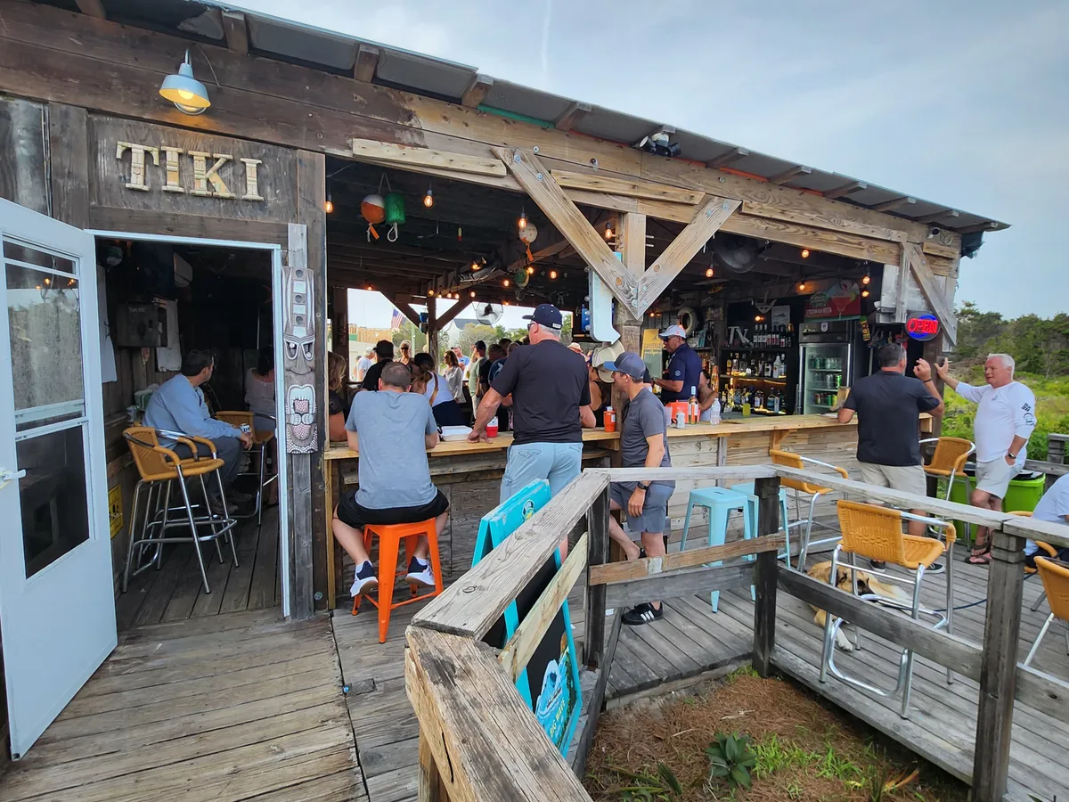 A vibrant exterior setting with a lively crowd gathered at a tiki bar, colorful seating, and a bar counter full of drinks. The atmosphere is festive, enhanced by sunset lighting, hinting at live music and local nightlife.