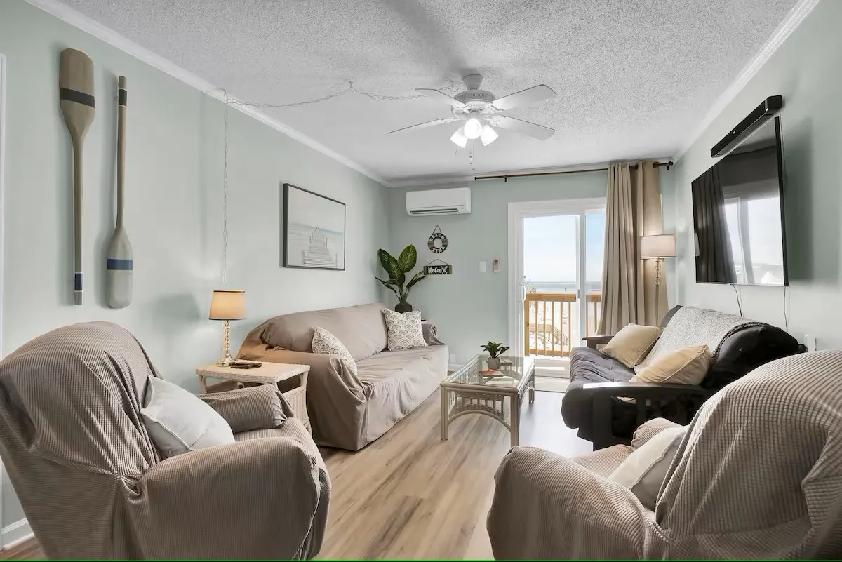 A cozy living room with a comfortable sofa area, nautical-themed oars mounted on the wall, and a TV. Floor-to-ceiling windows frame a stunning ocean view, seamlessly connecting the indoor space to the balcony and Atlantic scenery beyond.