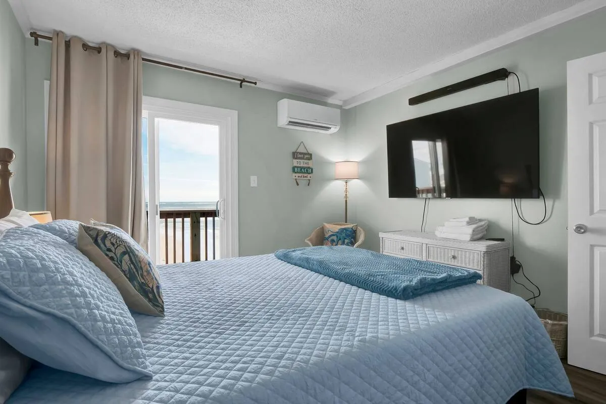 A tranquil bedroom featuring a bed with a blue quilt, a large TV mounted on the wall, and a window offering a serene ocean view. The decor is calm, with soft lighting that enhances the restful atmosphere, and the room feels spacious and inviting.