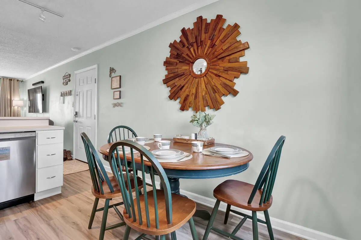 A cozy dining nook with a round table surrounded by green chairs, accented by a striking wooden sunburst mirror on the wall. The setting is ideal for shared meals, coffee chats, or working remotely in a comfortable setup.