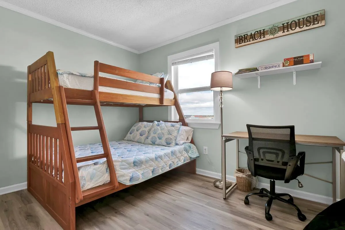A playful bedroom with a bunk bed, a desk and chair for studying or reading, a cheerful beach house sign, and a large window that lets in natural light. The space is inviting for families and children, blending fun with relaxation.