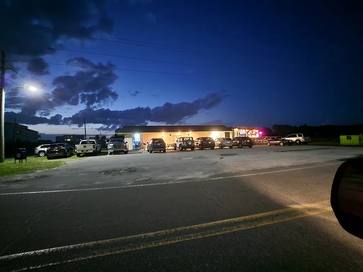 A warmly illuminated oceanfront building at dusk, with parked cars lining the front and streetlights casting a welcoming glow. The lively scene hints at communal gatherings, dining, and relaxed socializing just steps from the residence. Clouds drift overhead, adding to the serene, coastal atmosphere.