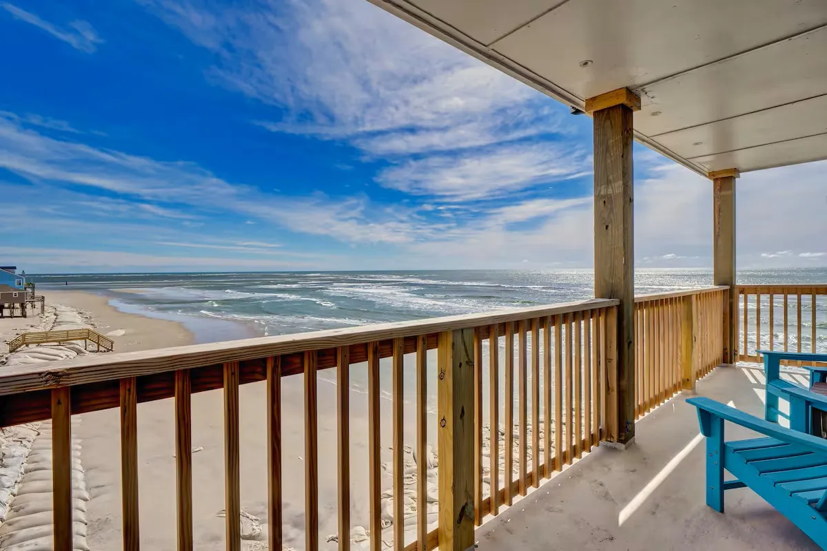 A serene oceanfront balcony with blue chairs arranged beside a wooden railing, overlooking rolling ocean waves and a sandy beach. The setting is ideal for sunrise coffees, sunset drinks, or quiet relaxation while taking in the view.
