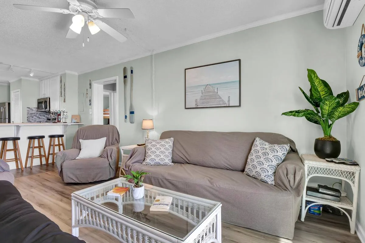 A bright, inviting living area featuring a comfortable sofa with patterned cushions, decorative wall art, an indoor plant, and bar stools lining the kitchen counter. The space feels both modern and cozy, ideal for family relaxation or casual gatherings.