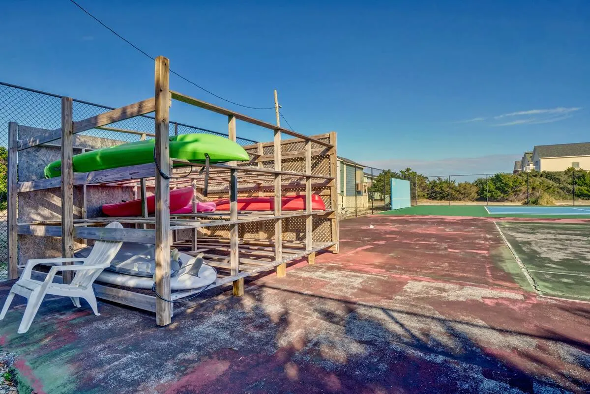 A row of brightly colored kayaks stacked near tennis courts, with a simple plastic chair nearby. The setting is playful and active, hinting at a range of outdoor recreational opportunities beyond just the beach.