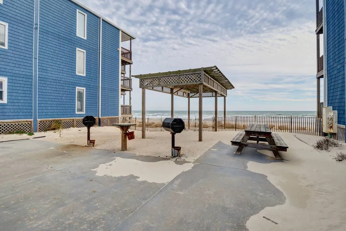 A casual outdoor area featuring blue seaside buildings, an inviting picnic table, a grilling area, and a clear view of the ocean beyond. The scene suggests a relaxed environment for outdoor dining, social gatherings, or simply soaking in the ocean breeze.