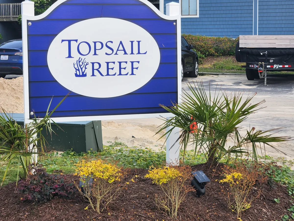 A welcoming exterior view featuring a landscaped sign, lush gardens, and the condo building in the background. The scene suggests arrival, relaxation, and the promise of memorable gatherings.