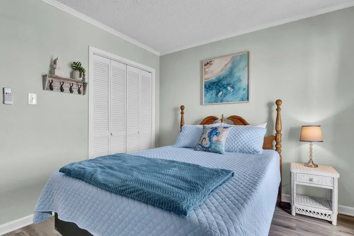 A sunlit bedroom with blue bedding, a decorative pillow, a nightstand topped with a lamp, and wall art above the bed. The room exudes peacefulness and intimacy, emphasizing comfort and thoughtful details for relaxation.