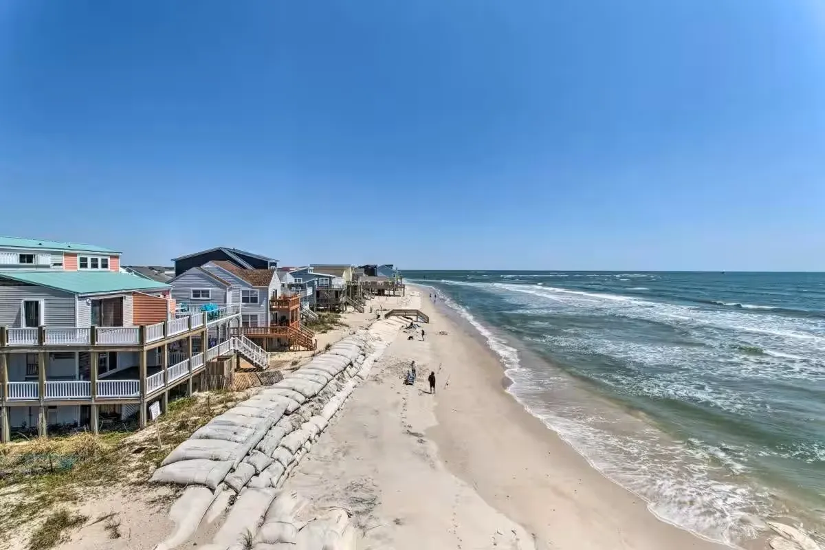 A tranquil beach scene with soft sand dunes, gently crashing waves, beachfront houses, and people taking a relaxed walk along the shore. The lighting suggests either early morning or sunset, lending a serene, inviting atmosphere.